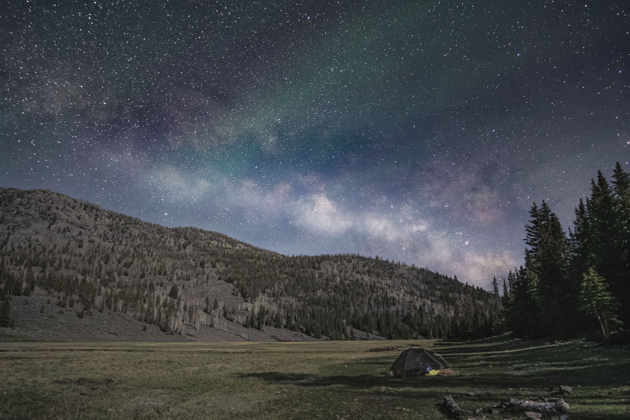 A tent sits in a valley, the milkey way stretches above, the sky is colored pink and green in wafts